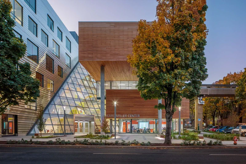 The Karl Miller Center at PSU at dusk — a timber-clad building with an angular glass atrium and KARL MILLER CENTER signage, fronted by trees on SW Broadway.